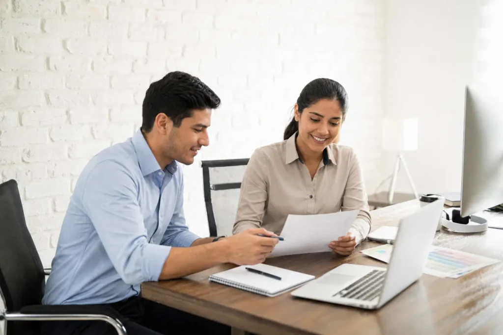 Two colleagues reviewing paperwork beside a laptop.