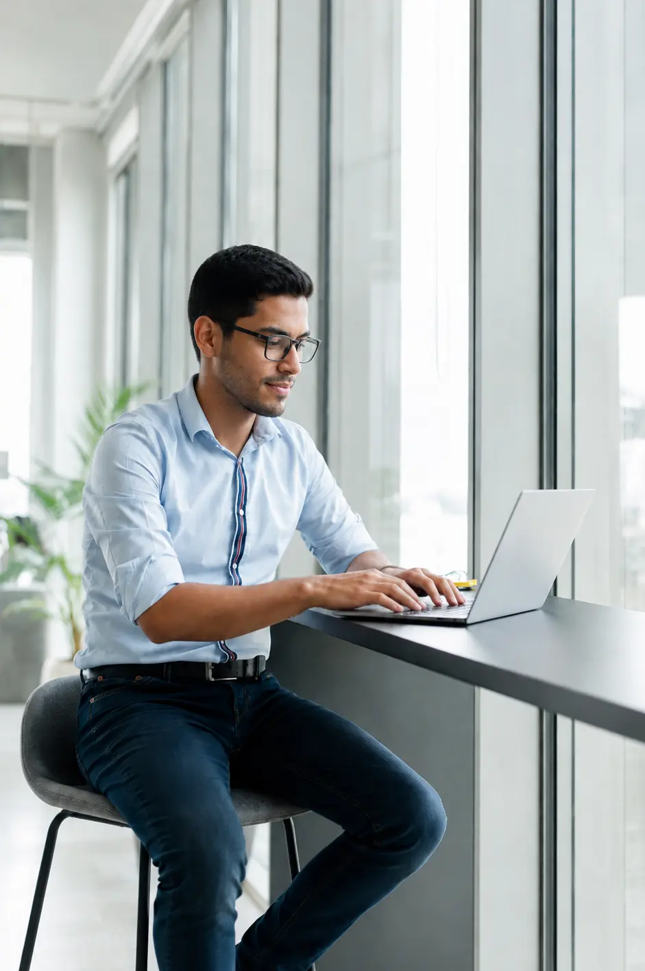 Man using a laptop while seated beside a large window.