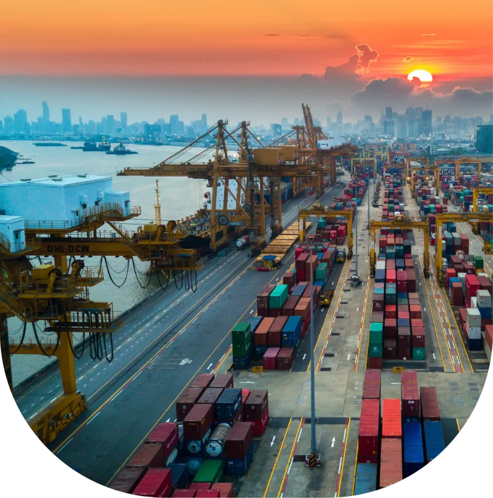 Shipping containers, cargo cranes, and vessels at a port at sunset.