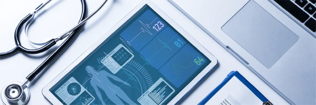 Stethoscope beside a tablet with medical icons and a laptop.