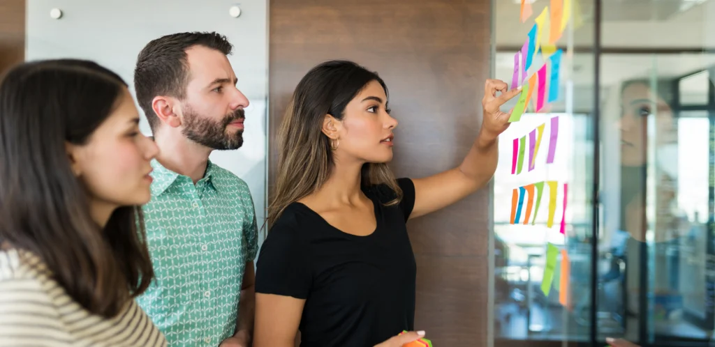 Three colleagues reviewing colorful sticky notes on a glass wall.