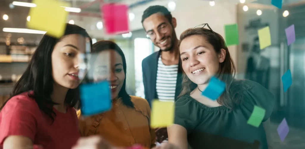 Four colleagues discussing colorful sticky notes on a glass wall.